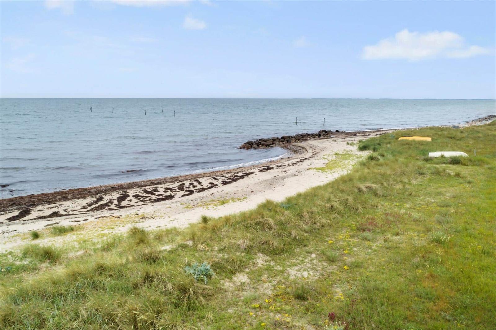 Strand mit Sand und Grasflächen, das Meer und ein kleiner Steinschutz sind sichtbar.