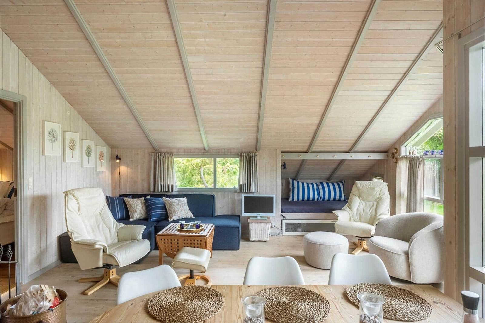 Wohnraum mit Holzdecke, Sofa, Sessel und Essbereich. Fenster mit Blick auf Grün.