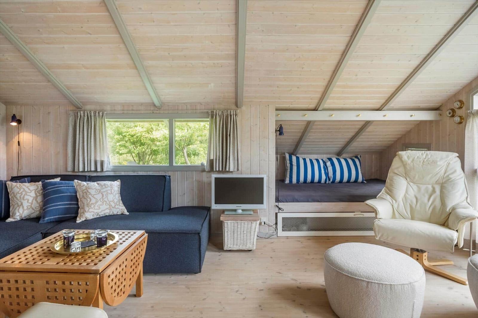 Wohnraum mit Sofa, Schlafcouch, Sessel und TV. Holzdecke und Fenster mit Blick auf Bäume.