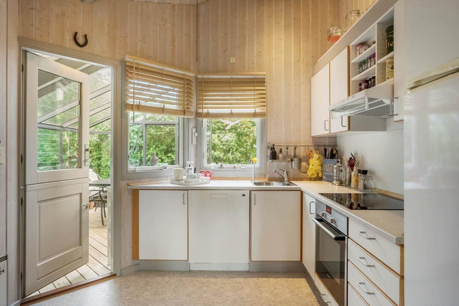 Küche mit Holzwänden, Fenstern und Tür zum Garten. Weiße Schränke und Küchengeräte.