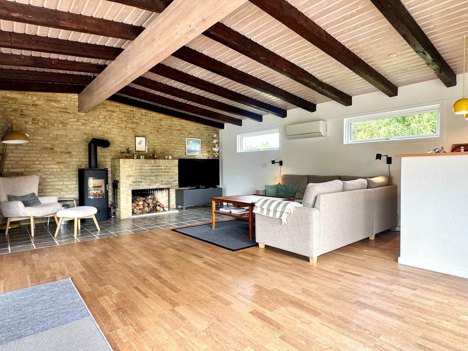 Wohnzimmer mit Holzboden, Kamin und Sofa. Deckenbalken und Fenster sichtbar.