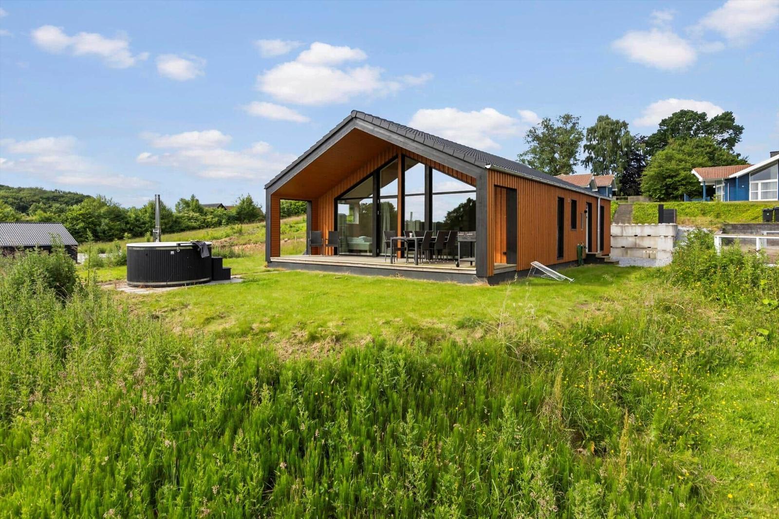 Modern wooden cabin with large glass area and outdoor space with lounge chairs.