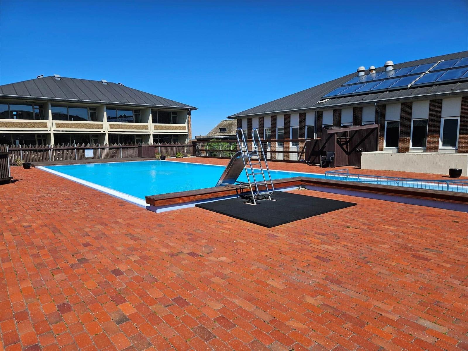Pool mit Rutsche und Treppe auf rotem Pflaster. Hintergrund: Gebäude mit Solarzellen.