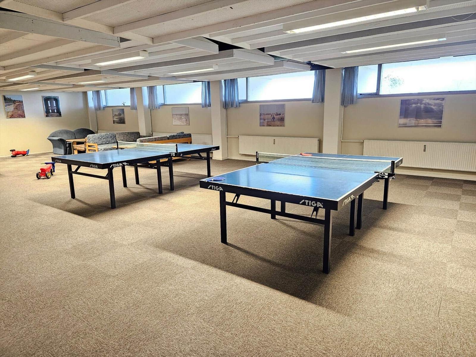 Zwei Tischtennistische in einem Raum mit Fenstern und Sofas.