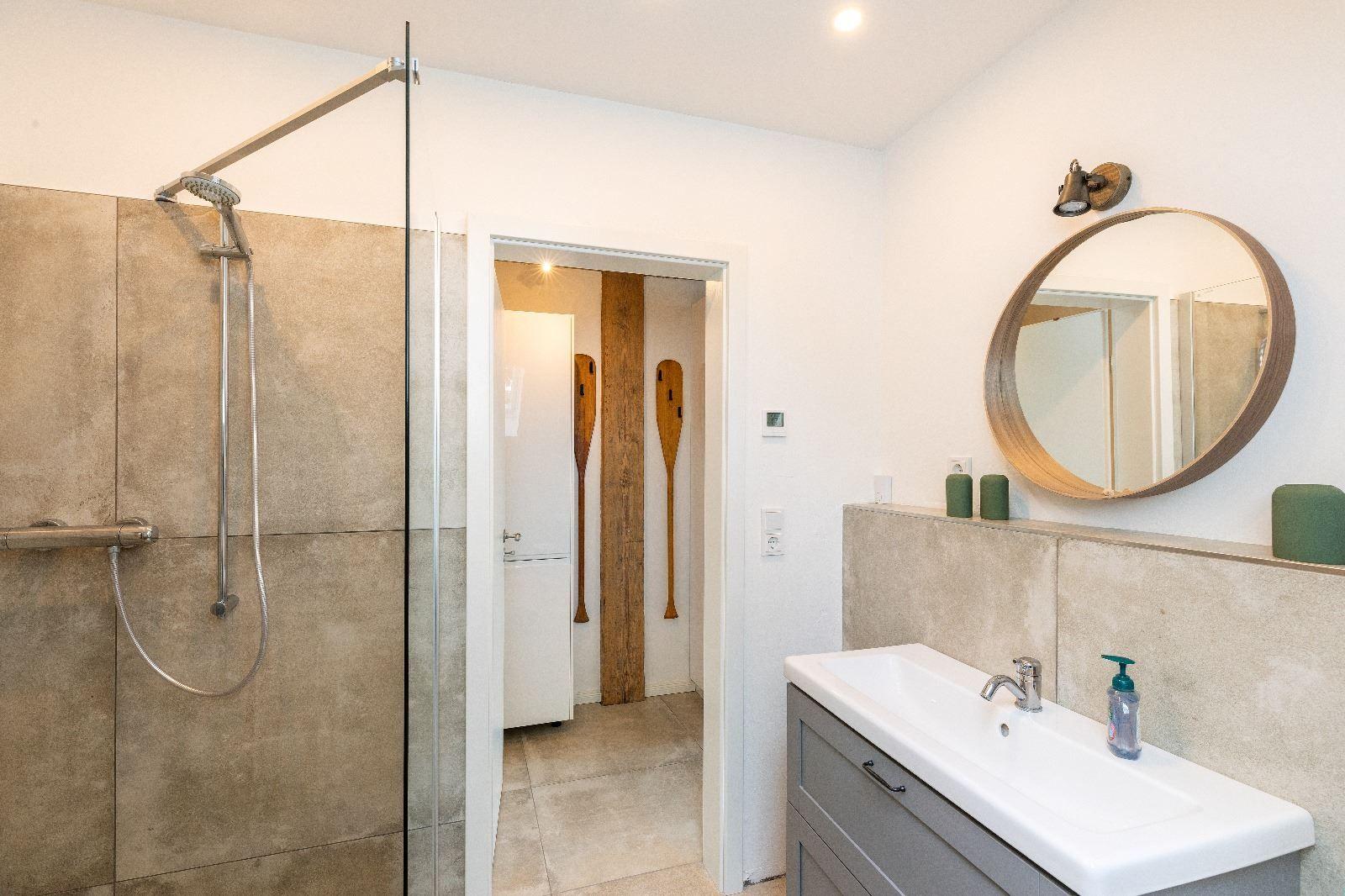 Badezimmer mit Dusche, Waschbecken und runden Spiegel. Holz-Ruder an der Wand.