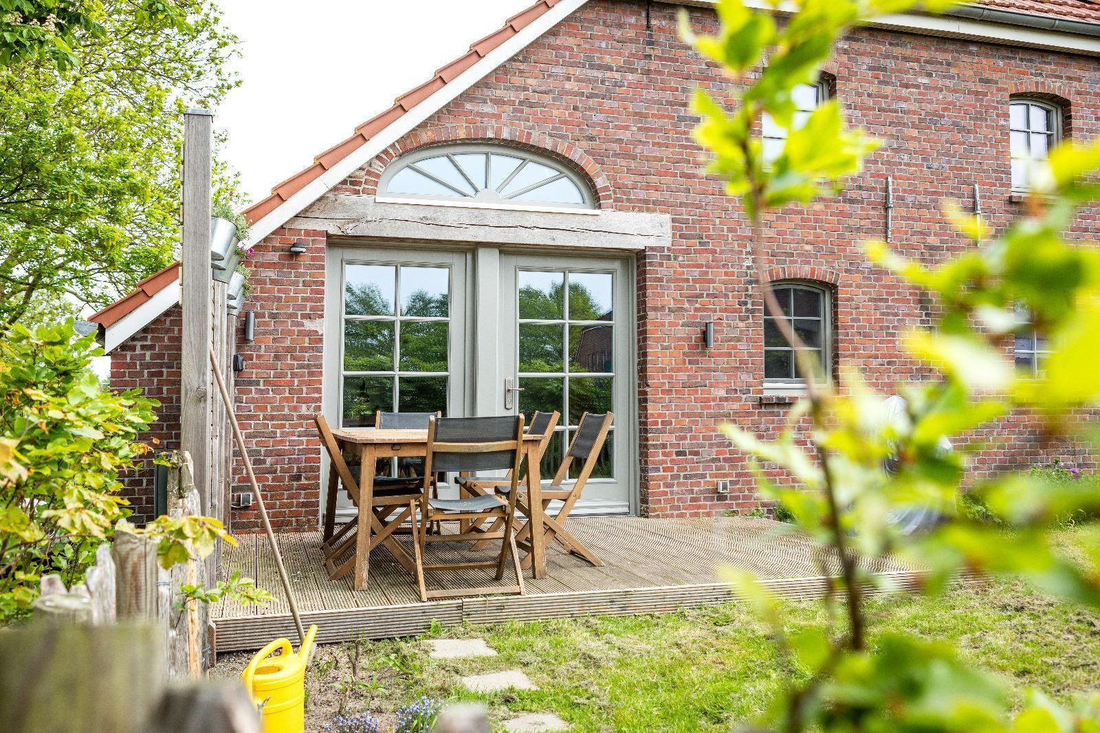 Roter Backsteinbau mit Terrasse und Holztisch. Garten mit Gras und Bäumen.