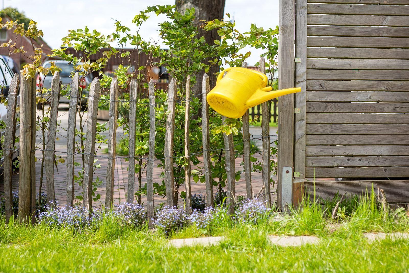 Gelber Gießkanister hängt an einem Holzzaun neben einem Garten mit Gras und Blumen.