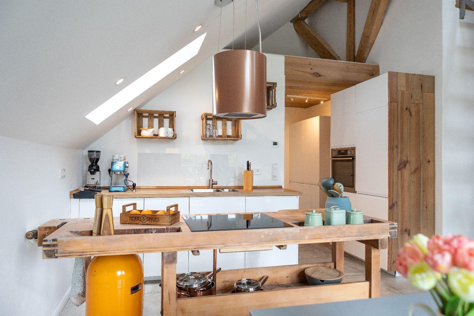 Küche mit Holzinsel, weißen Schränken und Dachfenster. Holzkonstruktion und Küchenutensilien sichtbar.