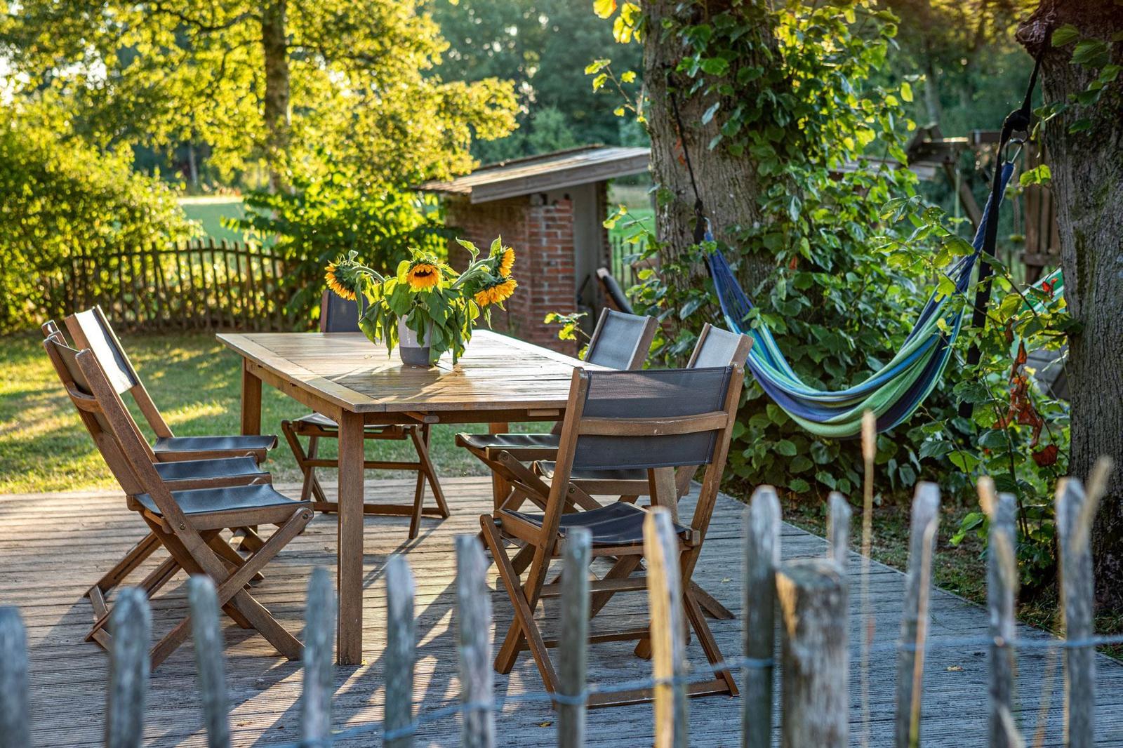 Holztisch mit Stühlen und Sonnenblumen auf Terrasse. Hängemattensitz unter Baum.