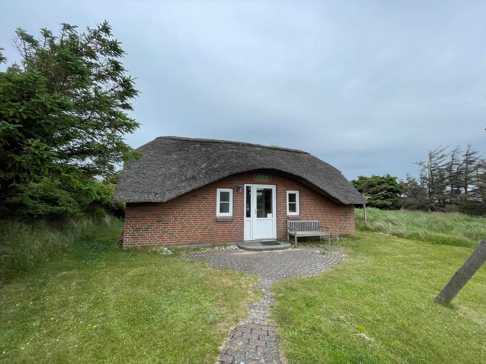 Ein kleines Haus mit Strohdach, rotem Backstein, weißer Tür und Bank auf dem Vorgarten.