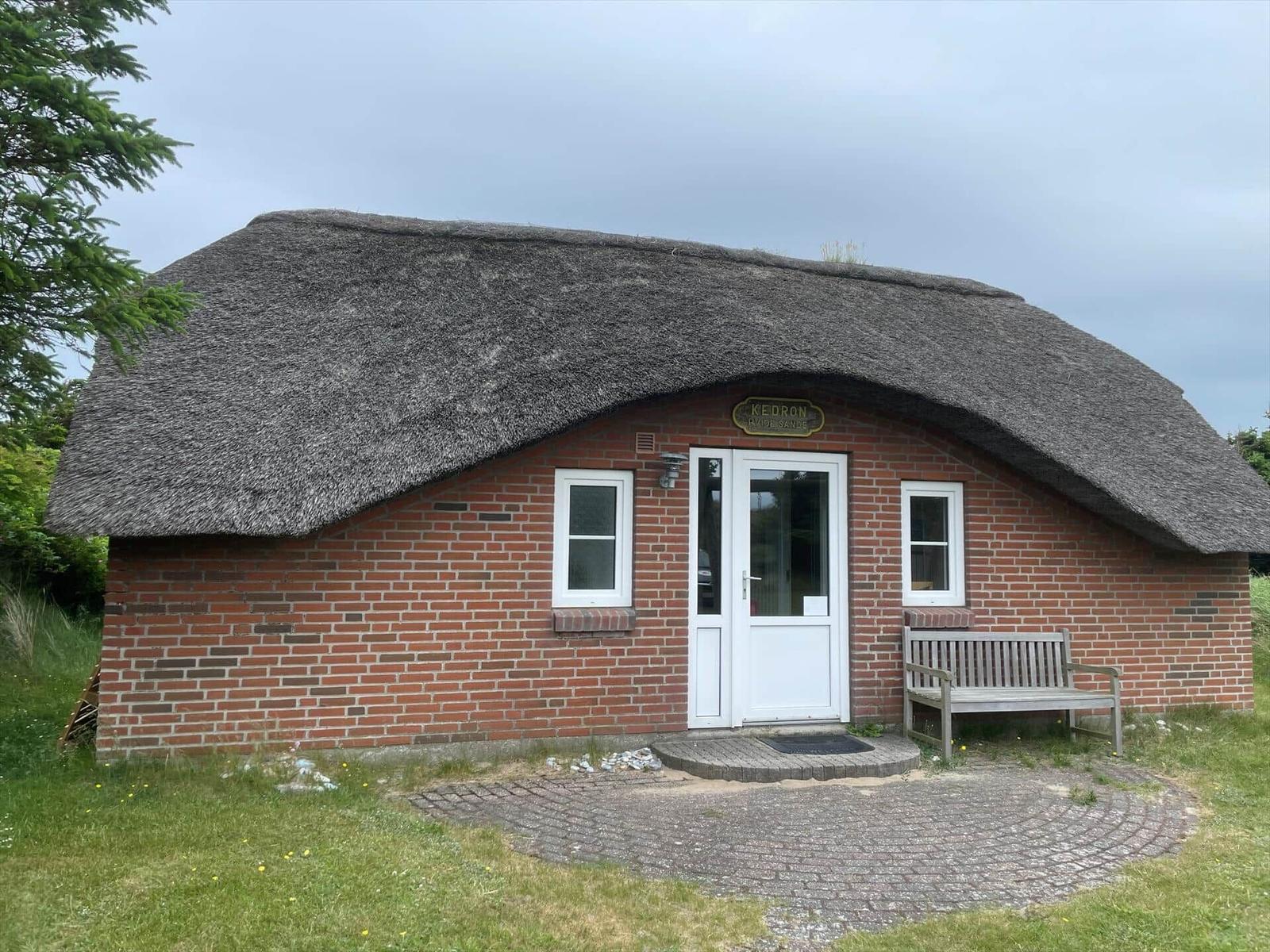 Haus mit Strohdach, roten Ziegeln, weißer Tür und Bank vor dem Eingang.