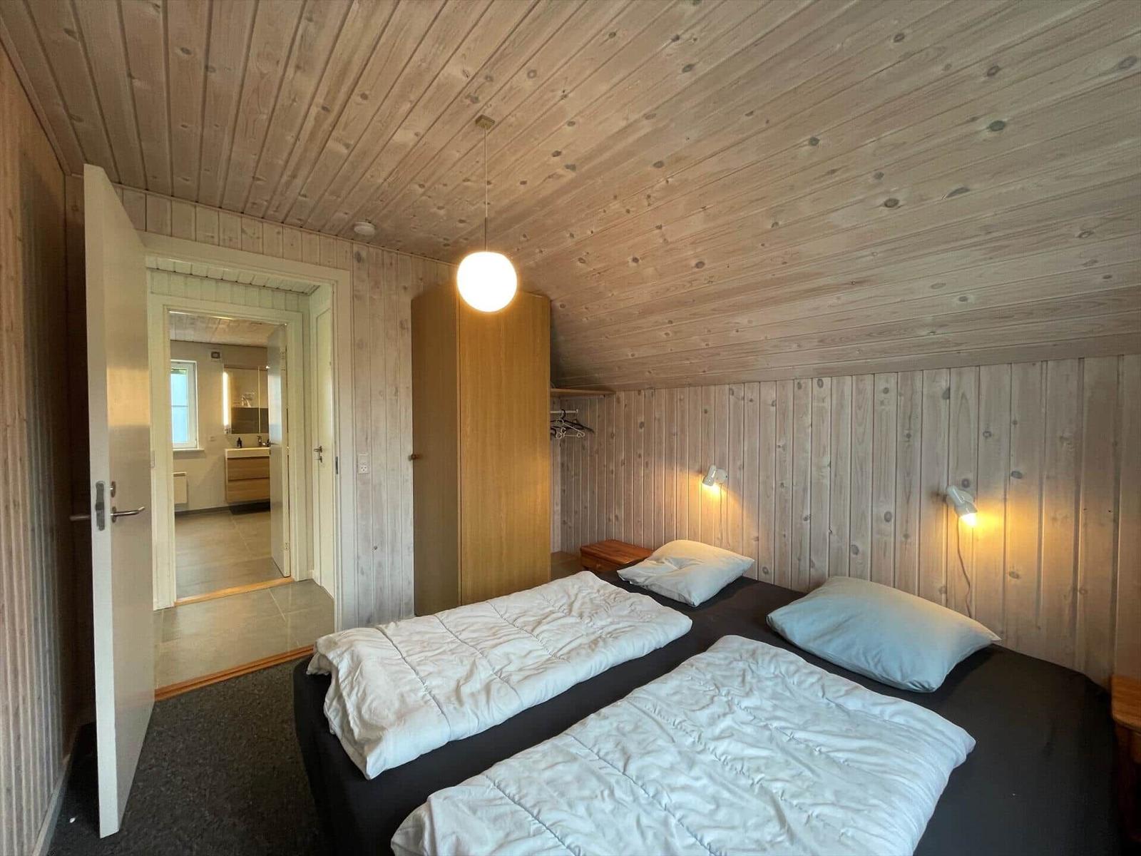 Zimmer mit zwei Betten, Holzwand und -decke, Nachttische und Lampen, Tür zum Bad.