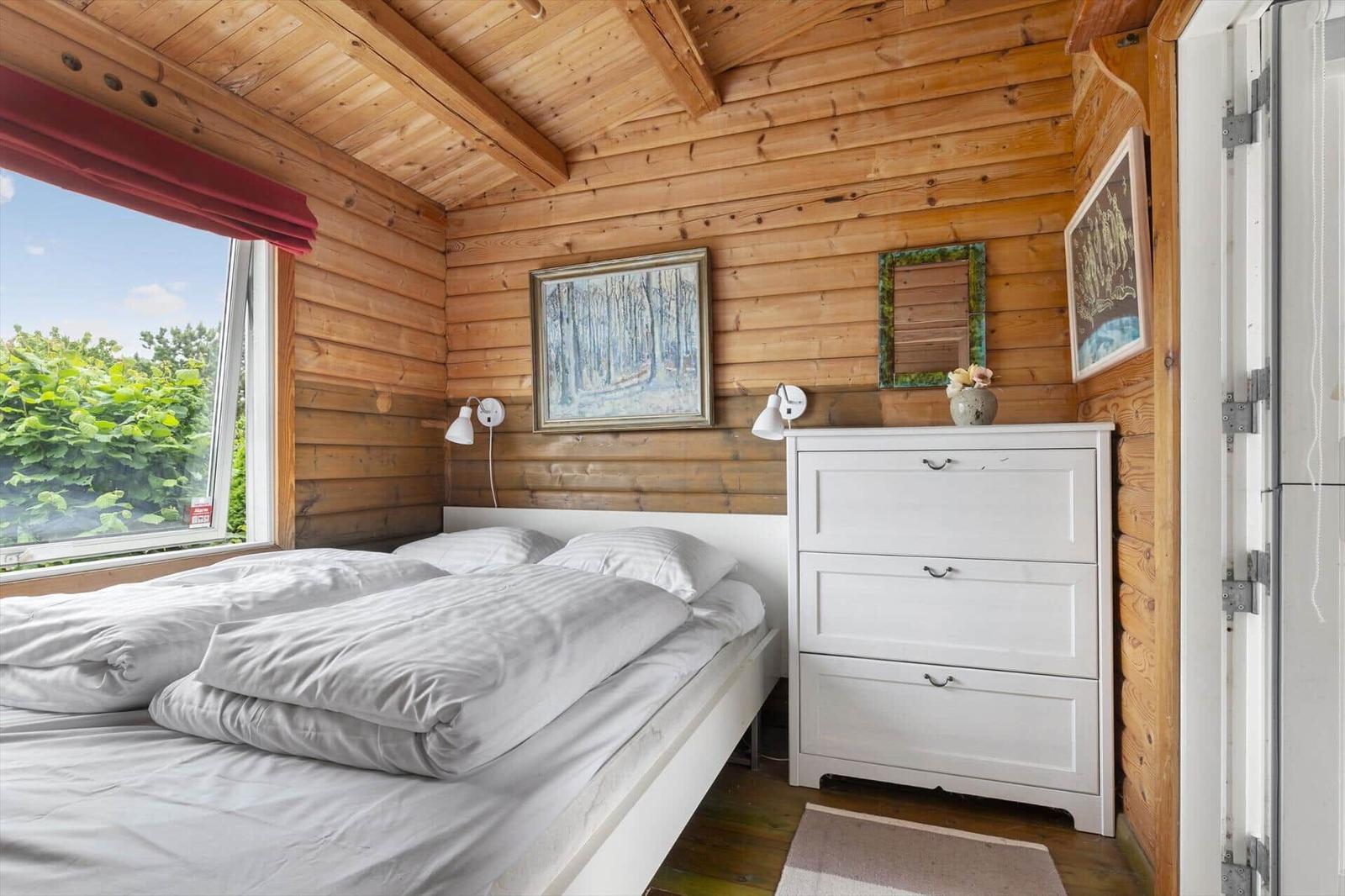 Schlafzimmer mit Holzwand, weißem Bett und Schrank. Fenster mit Blick auf grüne Vegetation.