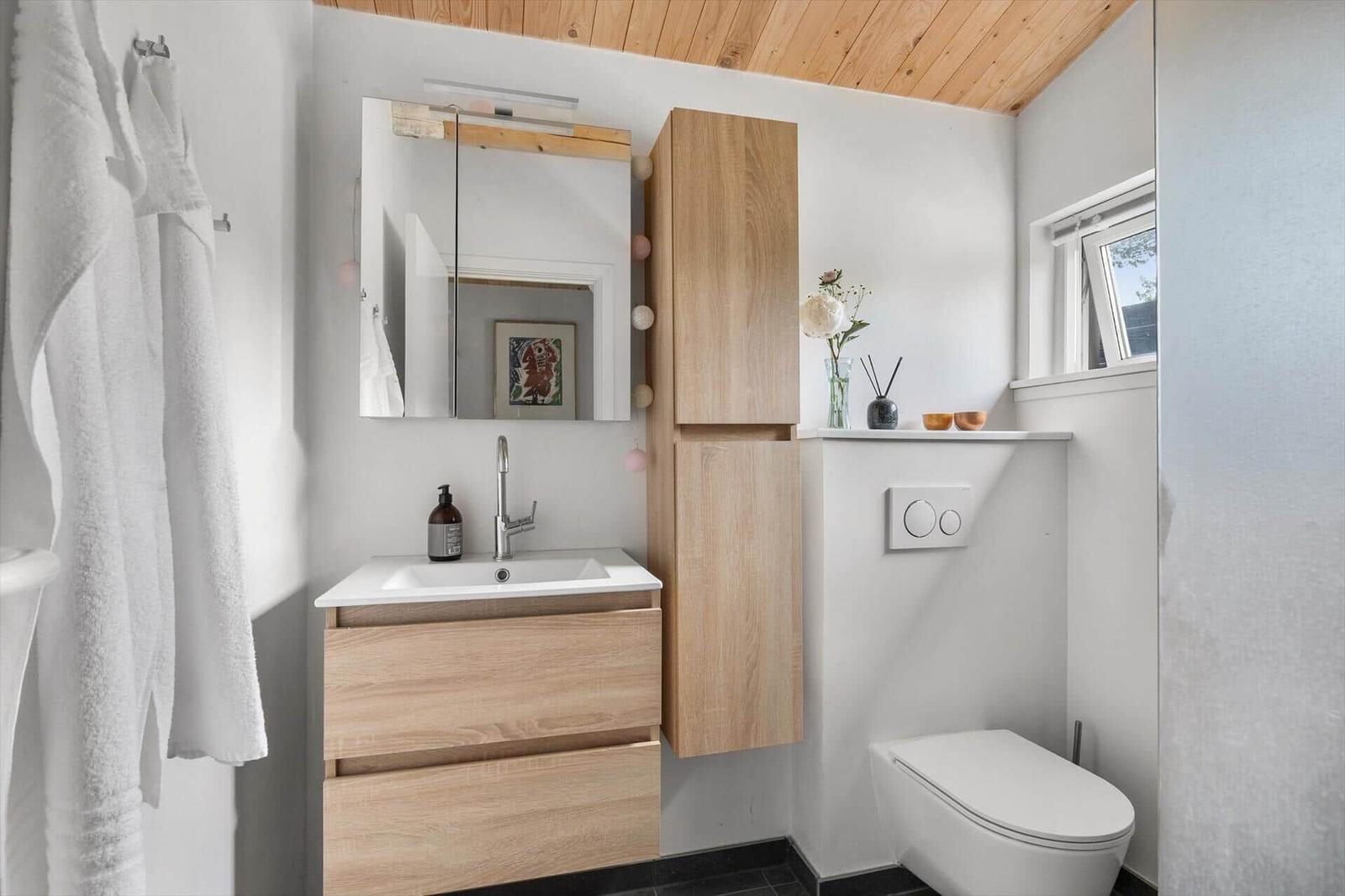 Badezimmer mit Holzregalen, Waschbecken und Toilette. Fenster und Spiegel sichtbar.
