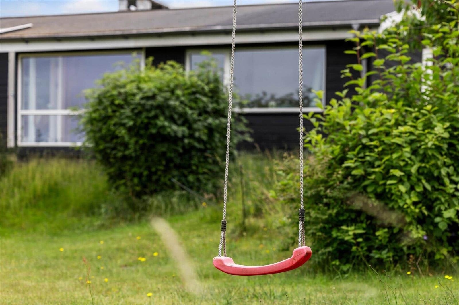 Ein roter Schaukelstuhl hängt im Garten vor einem Haus mit grünen Sträuchern.