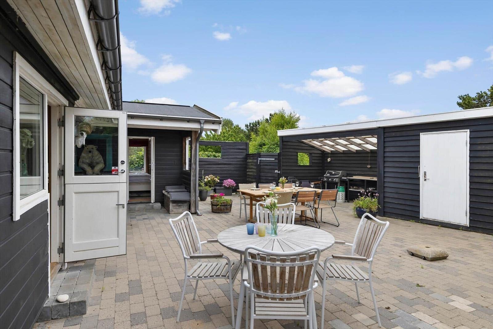 Terrasse mit Tisch, Stühlen und Grillbereich. Schwarze Holzwände und Pflanzen.