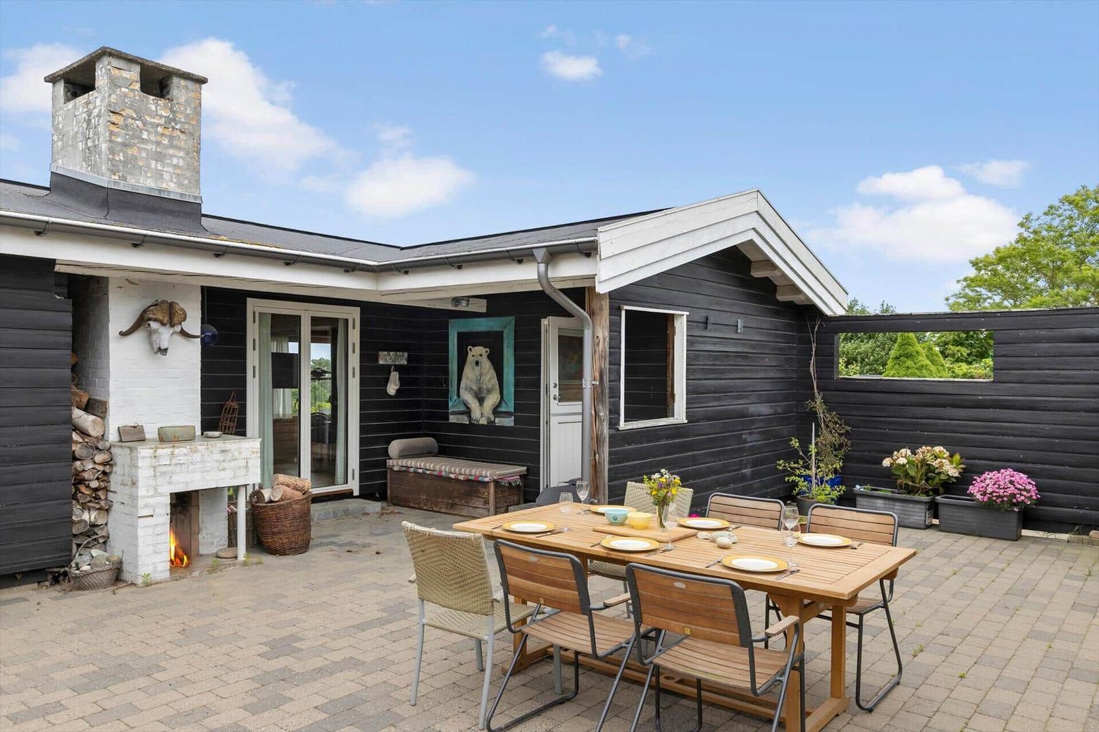 Terrasse mit Tisch, Stühlen und Grill. Schwarze Holzwände und weißer Kamin. Blumen und Pflanzen.