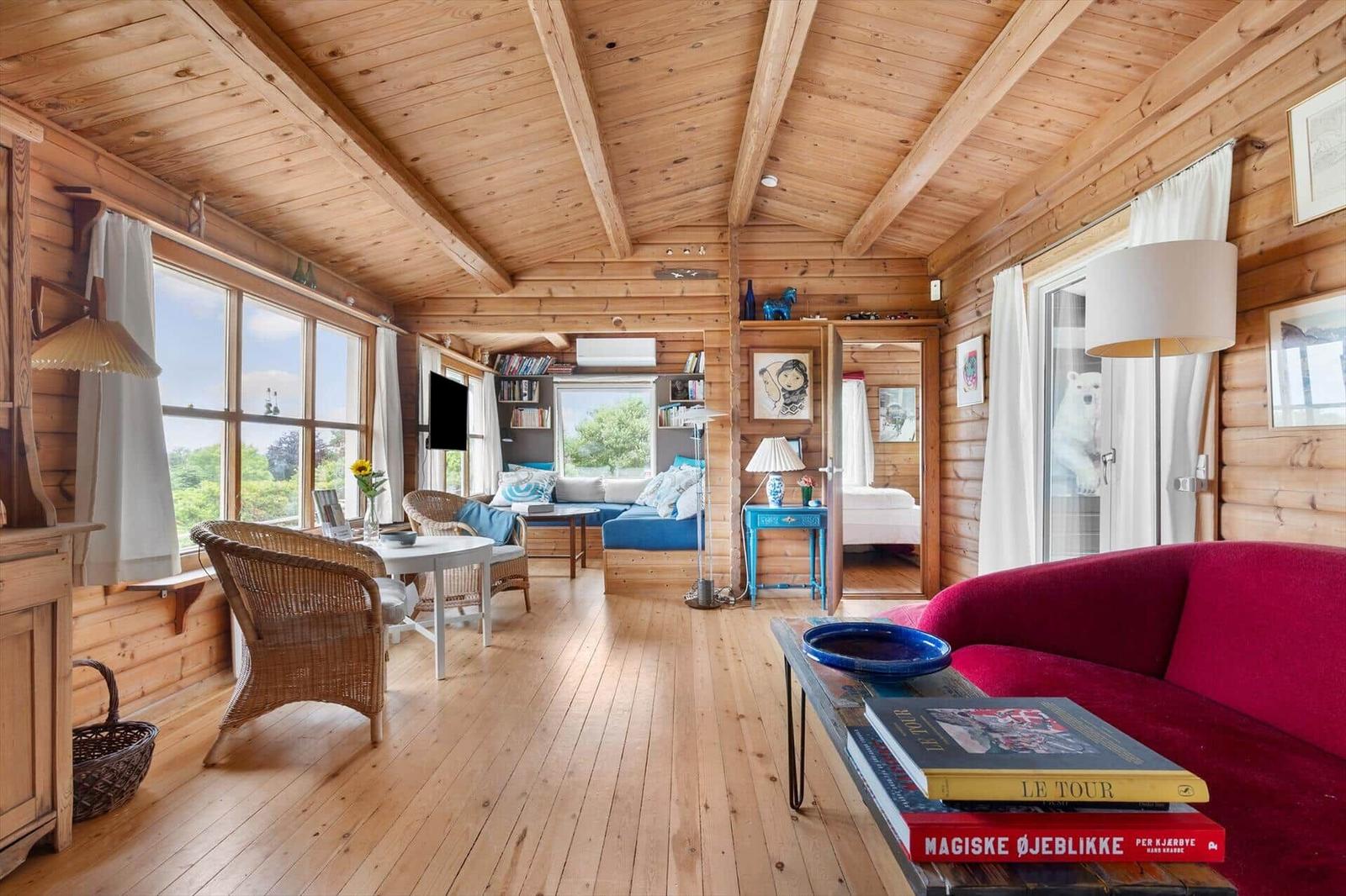 Wohnraum mit Holzdecke, Fenstern und rotem Sofa. Im Hintergrund ist ein Schlafzimmer sichtbar.