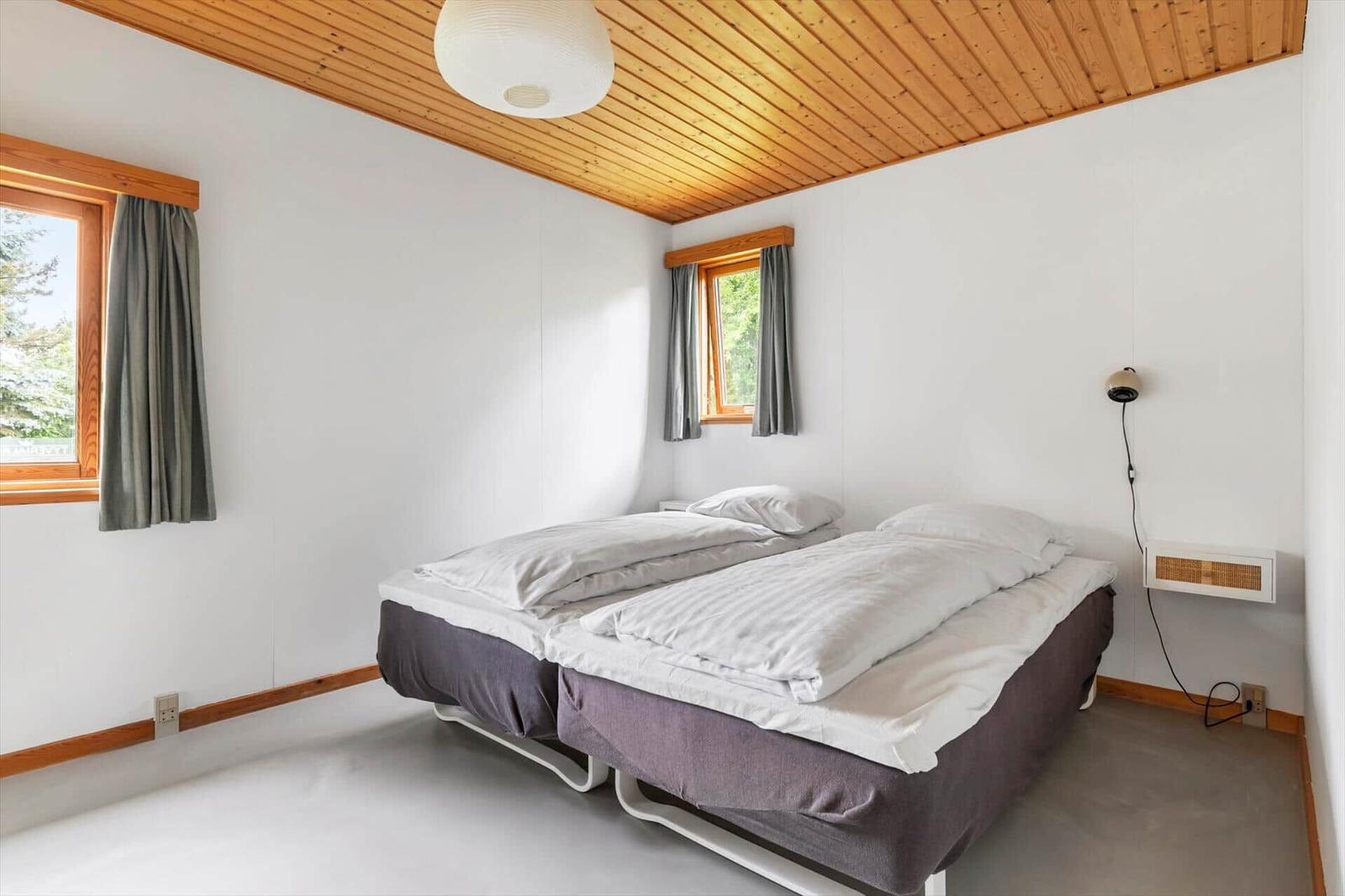 Two sleeping areas with bedding, wooden ceiling, and windows with curtains.