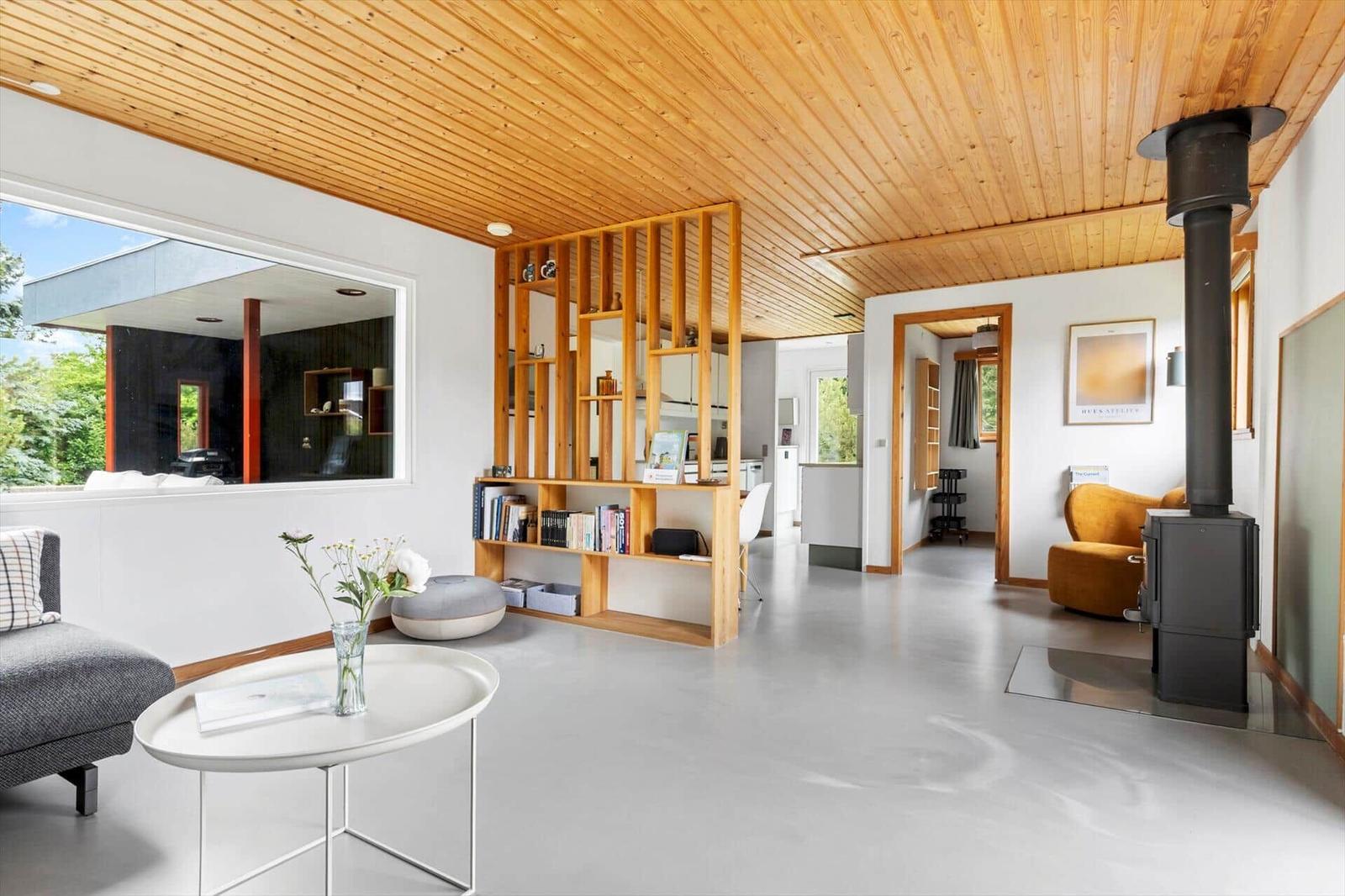 Living area with wooden ceiling, fireplace, and large window. Bookshelf and seating areas.