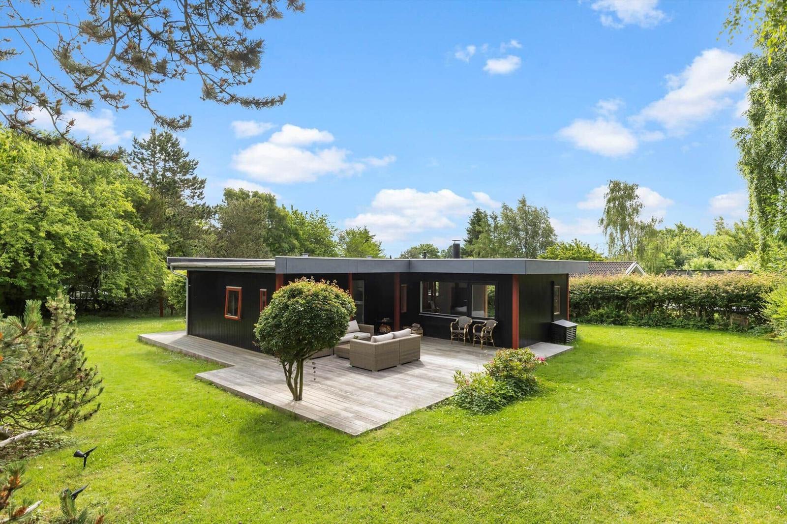 Modern black cabin with wooden terrace and garden. Outdoor seating area under trees.