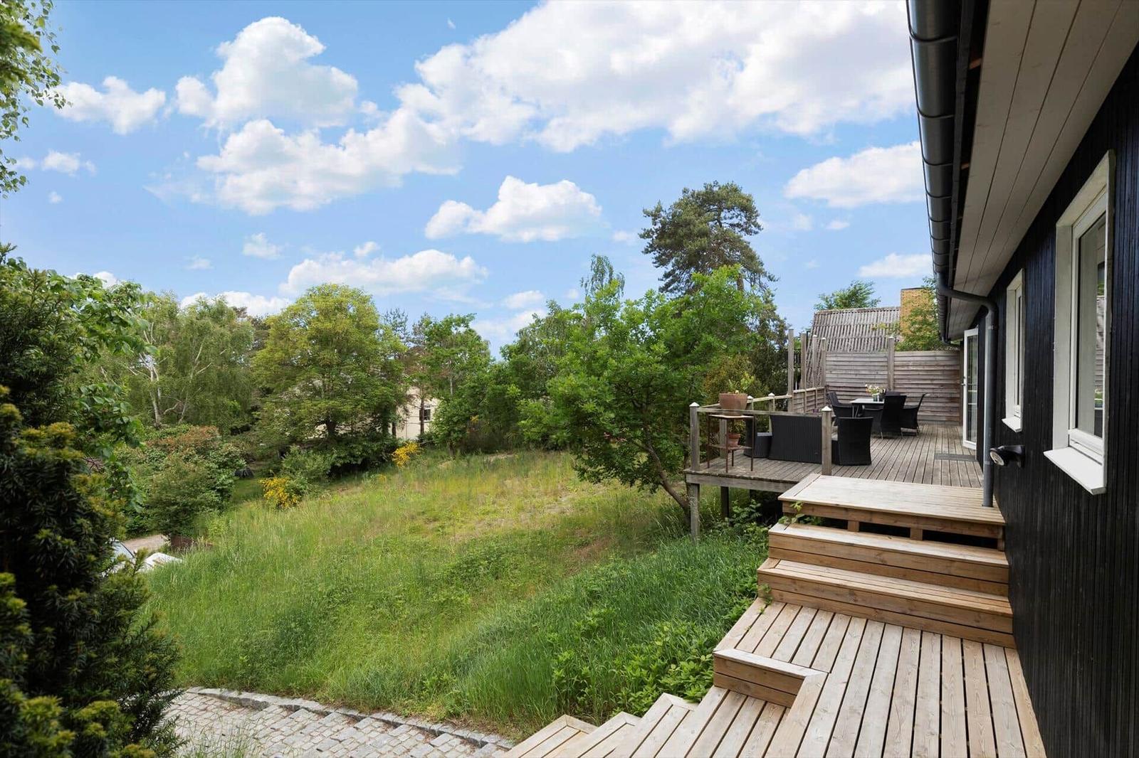 Wooden terrace with seating, view of green garden and trees under blue sky.