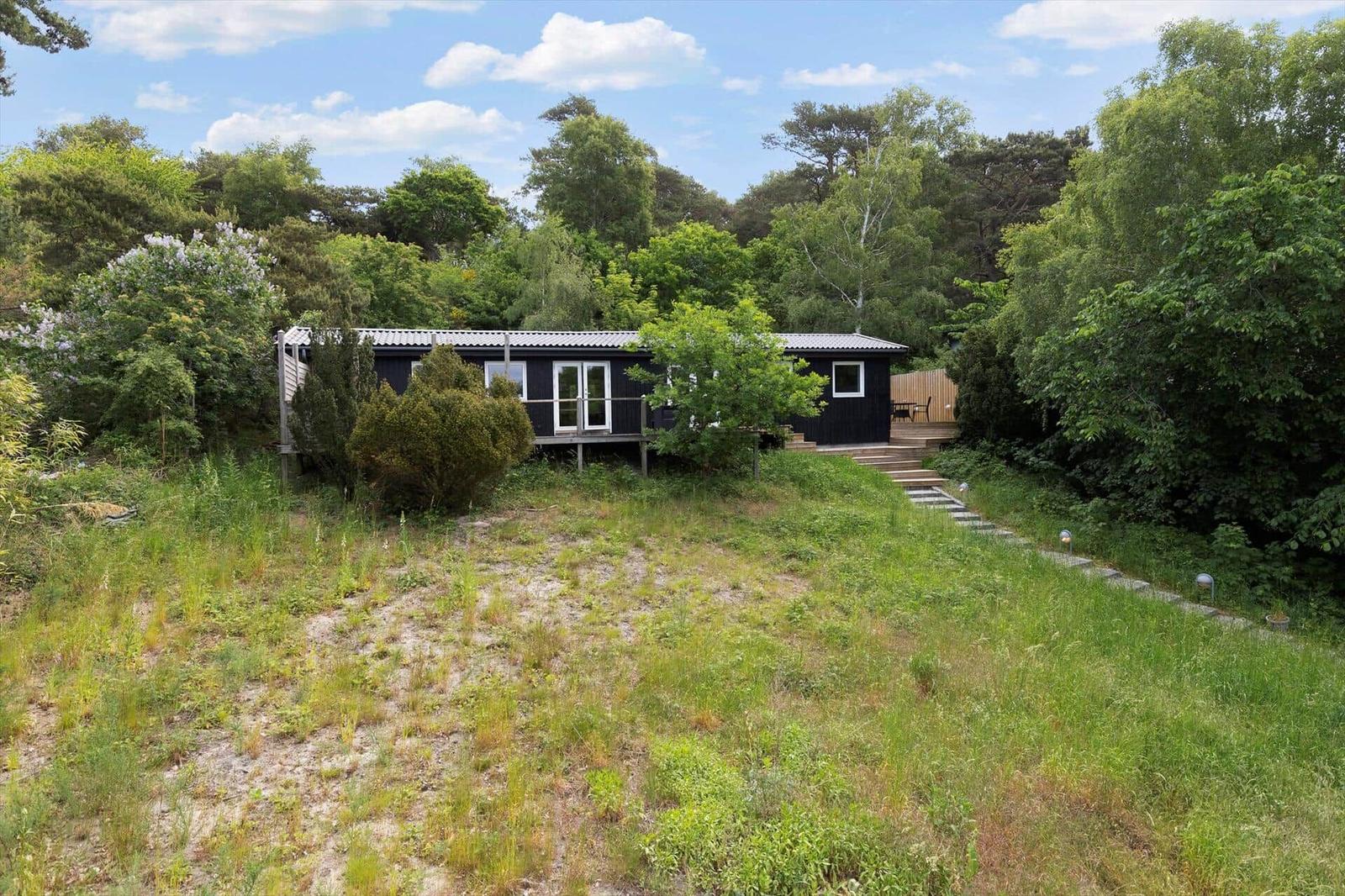 Black wooden house on stilts, surrounded by trees and grassy areas with stone steps.