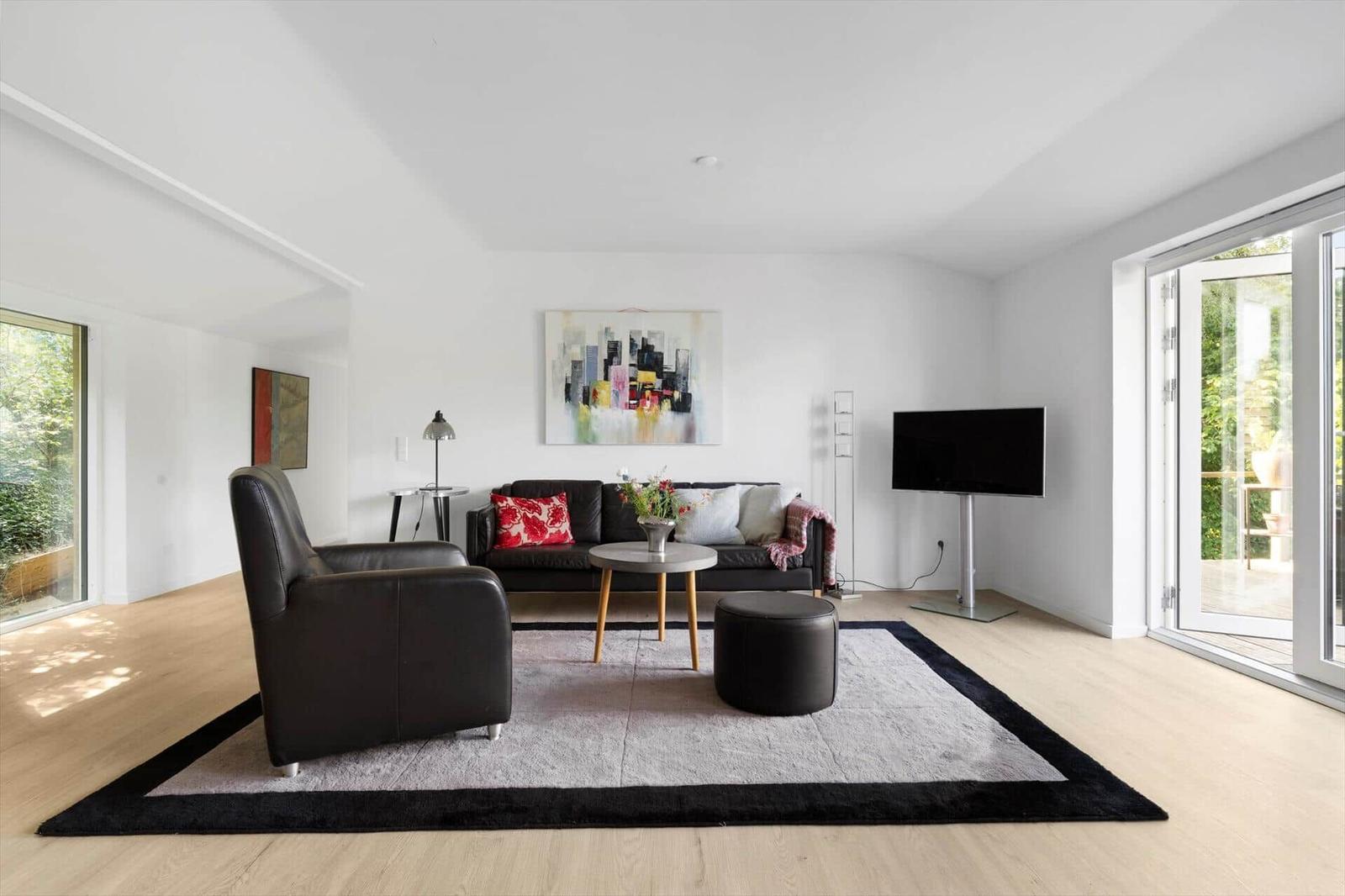 Living room with sofa, armchair, rug, and TV. Large windows with garden view.