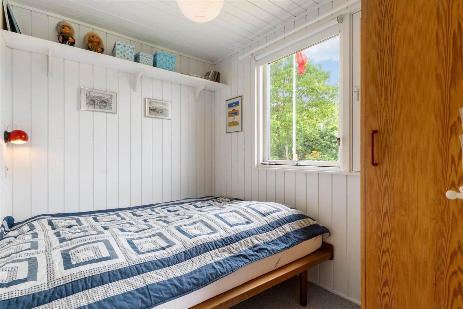 Schlafzimmer mit Bett, Fenster und Holzschränkchen. Wandverkleidung weiß, Decke weiß.