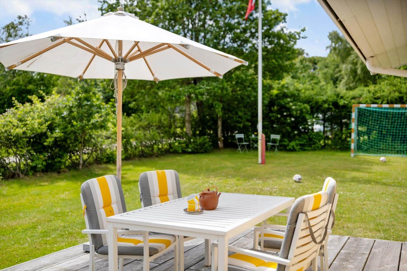 Terrasse mit Tisch, Stühlen und Sonnenschirm. Grasfläche mit Fußballtor im Hintergrund.
