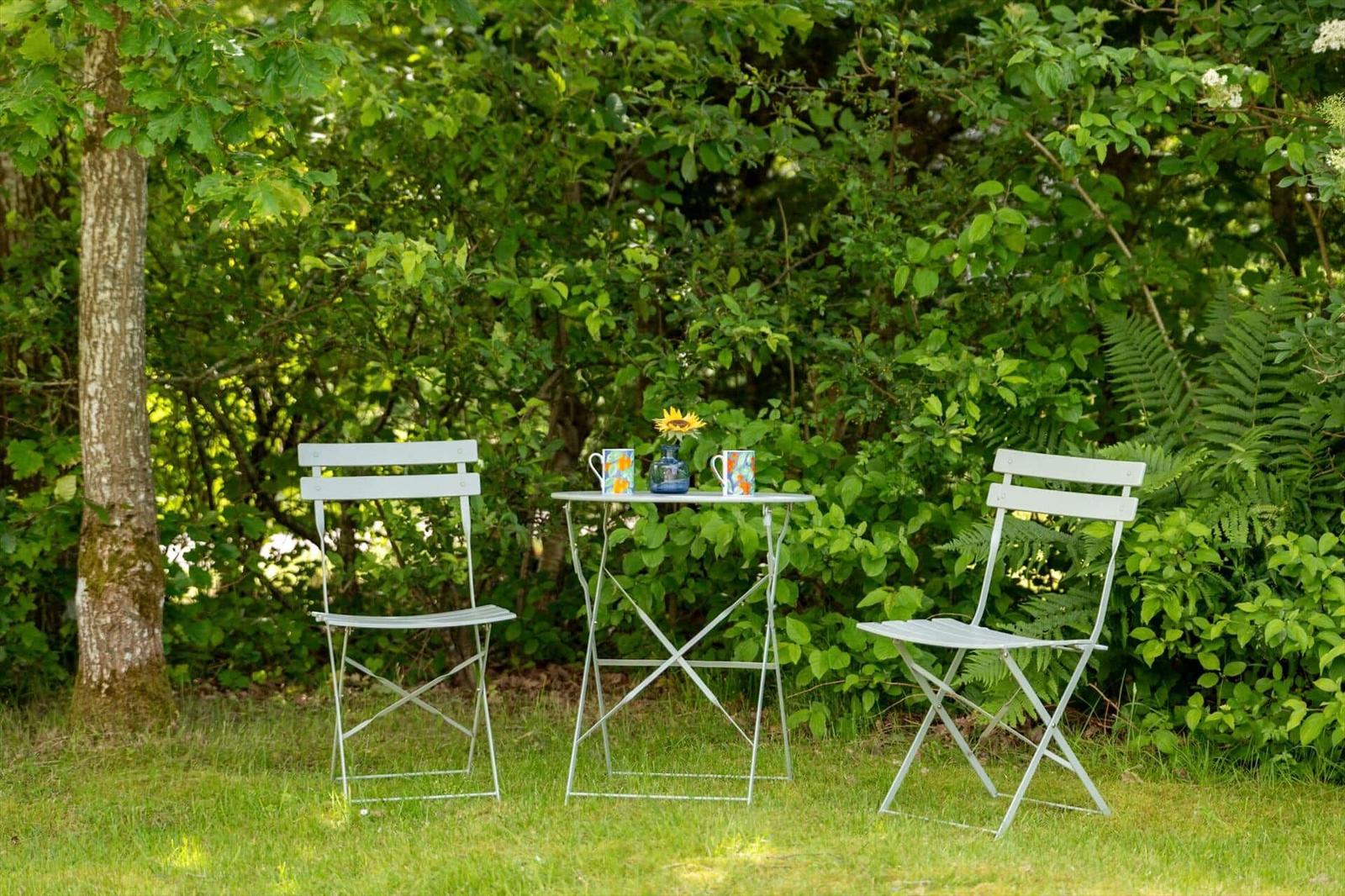Zwei weiße Stühle und ein Tisch stehen im Gras vor dichtem Grün.
