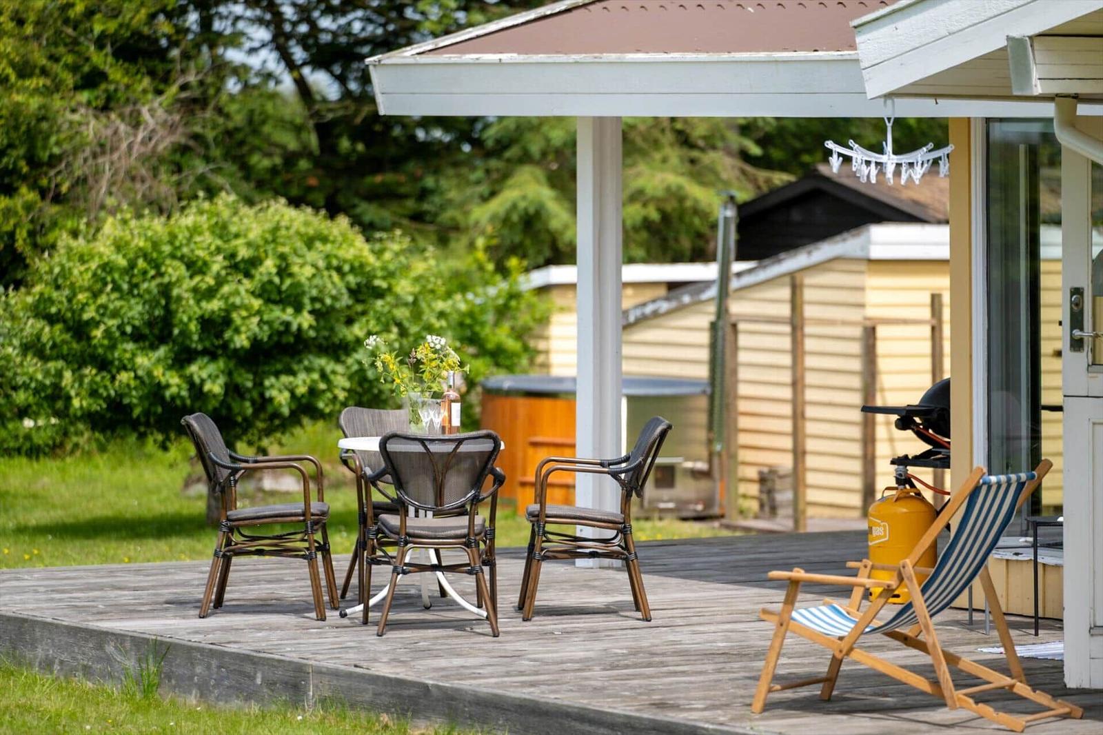 Terrasse mit Tisch, Stühlen und Liegestuhl. Hintergrund: Grünfläche und Holzhaus.