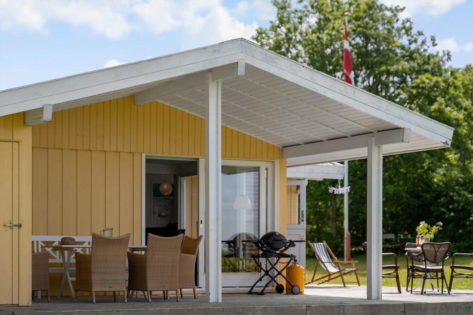 Terrasse mit Grill, Tischen und Stühlen unter weißem Dach. Daneben steht eine Liegestuhl.