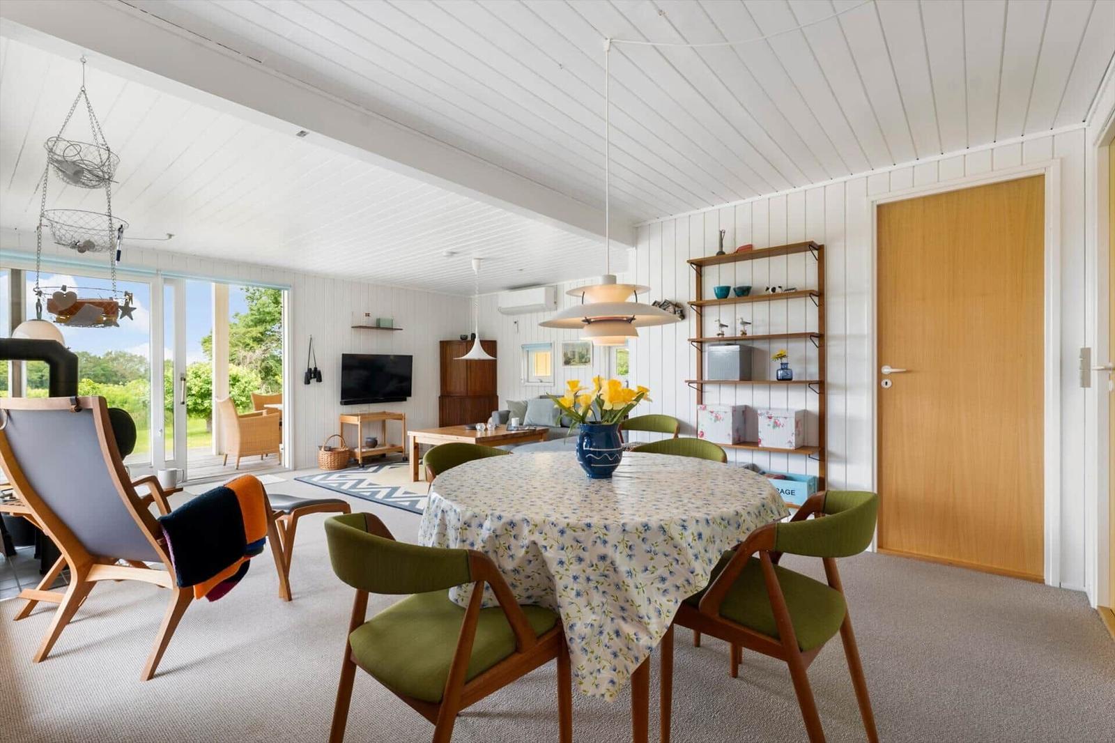Esszimmer mit runden Tisch und grünen Stühlen. Hintergrund: Wohnbereich mit TV und Terrasse.