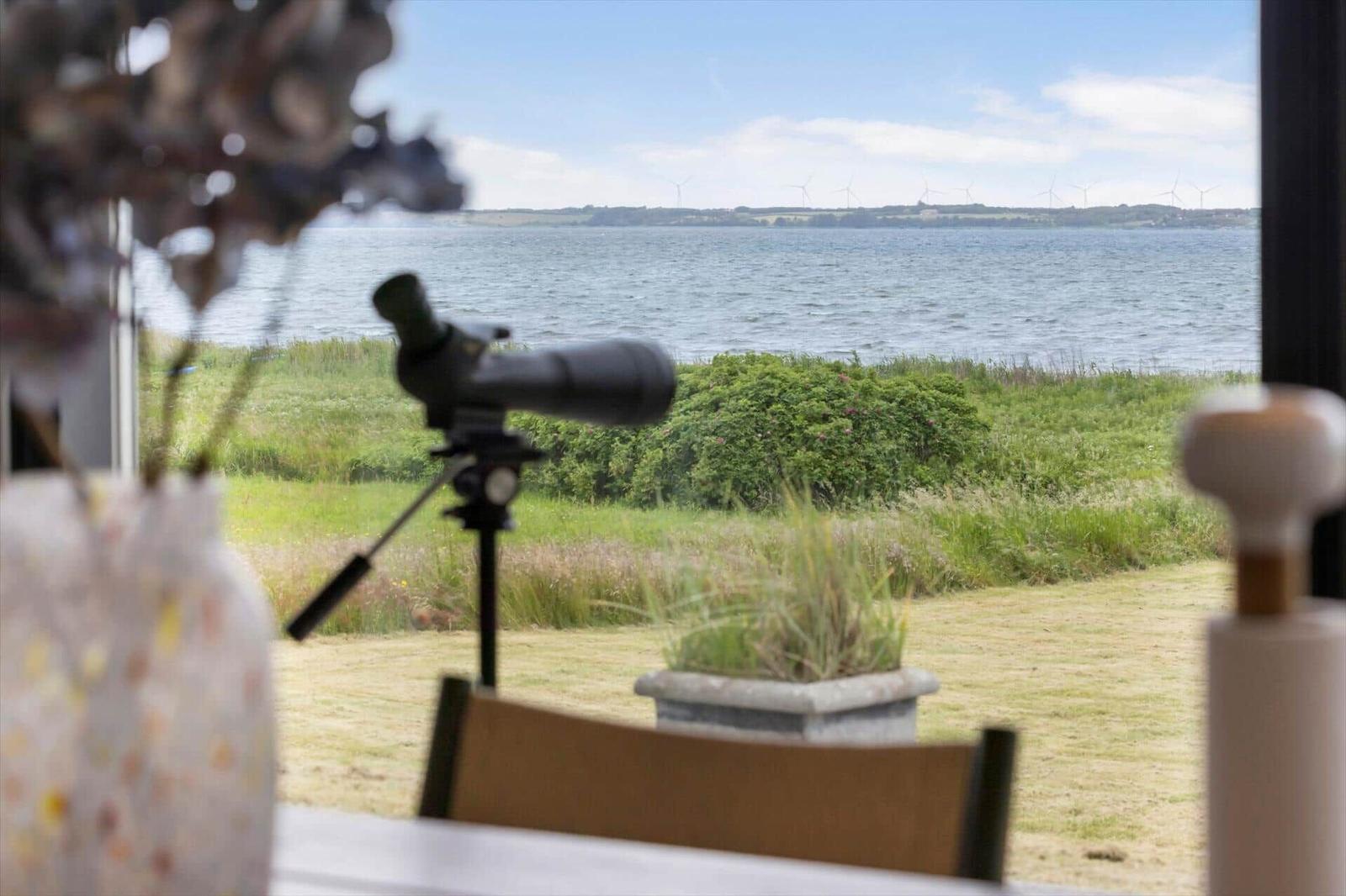 Durch das Fenster ist ein Blick aufs Wasser und Windräder zu sehen. Ein Fernglas steht auf einem Tisch.