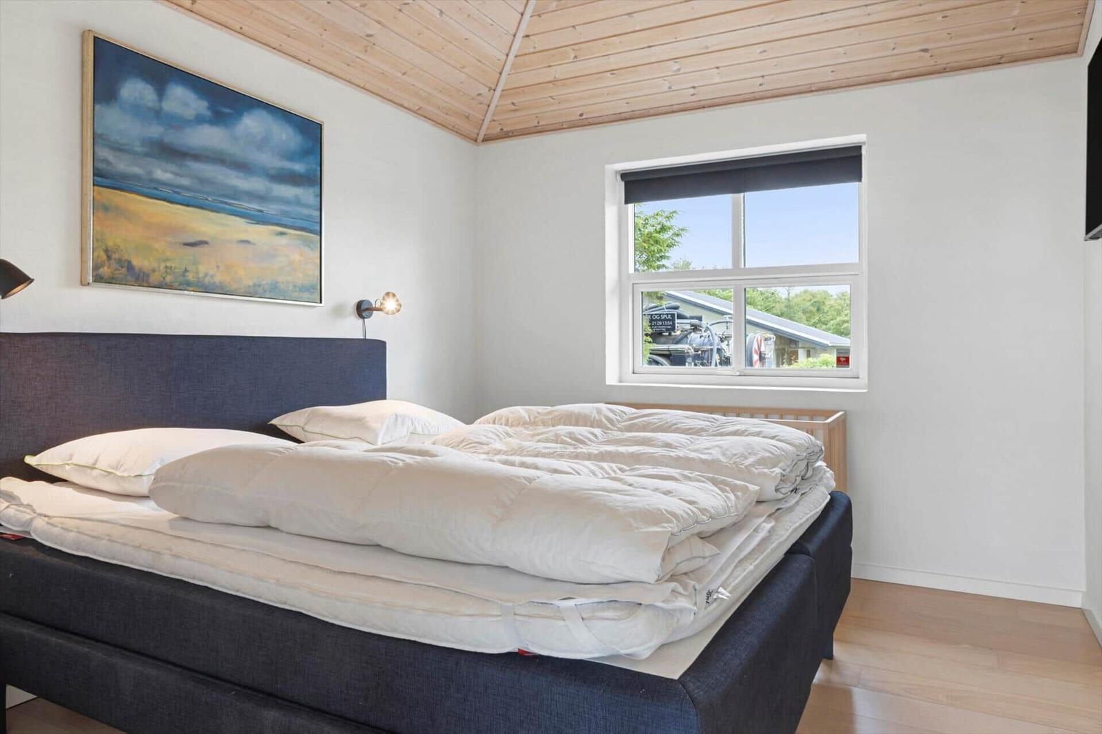 Schlafzimmer mit Bett, Wandbild und Fenster. Holzdecke und hellbrauner Boden.