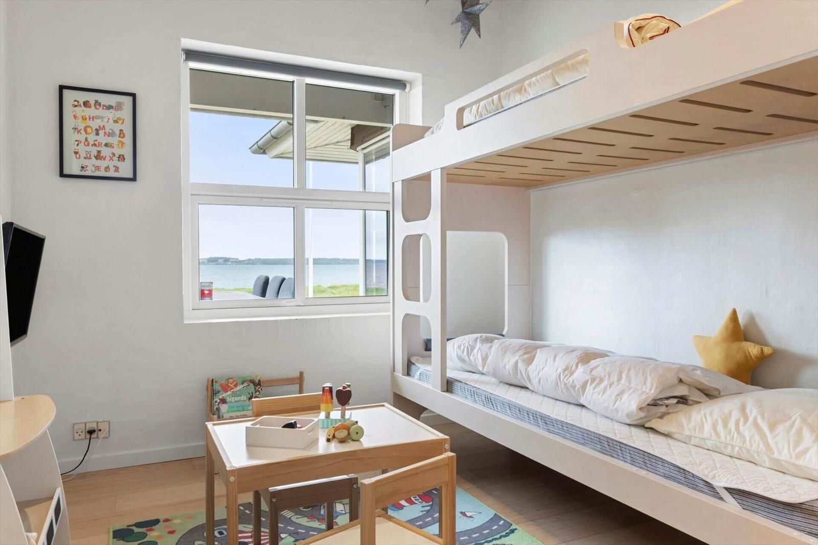 Kinderzimmer mit Doppelbett, Tisch und Fensterblick auf Wasser.