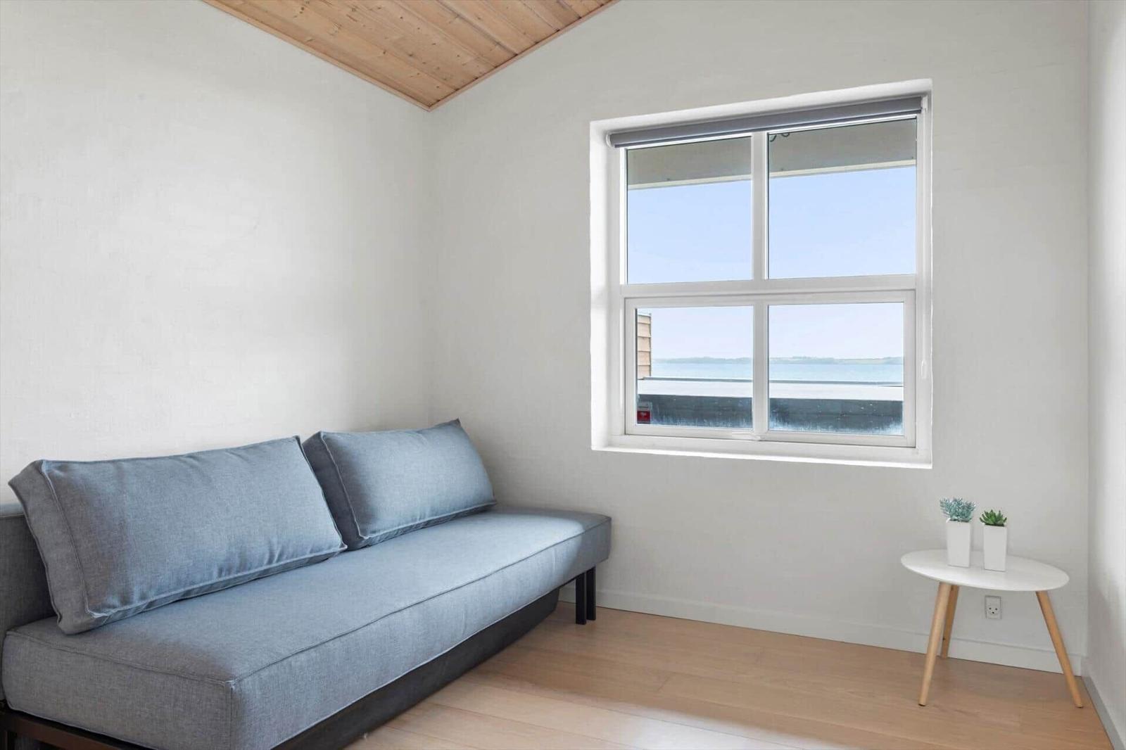 Ein Raum mit einem grauen Sofa, einem Fenster mit Blick auf Wasser und einem kleinen Tisch mit Pflanzen.