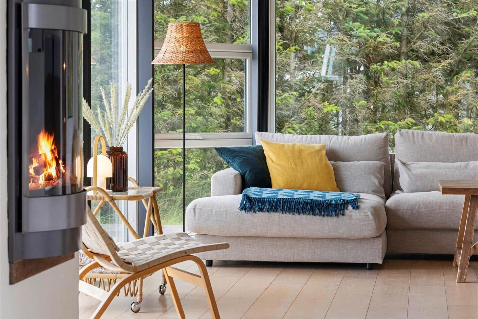 Ein Wohnzimmer mit Sofa, Kamin und großen Fenstern. Draußen sind Bäume sichtbar.