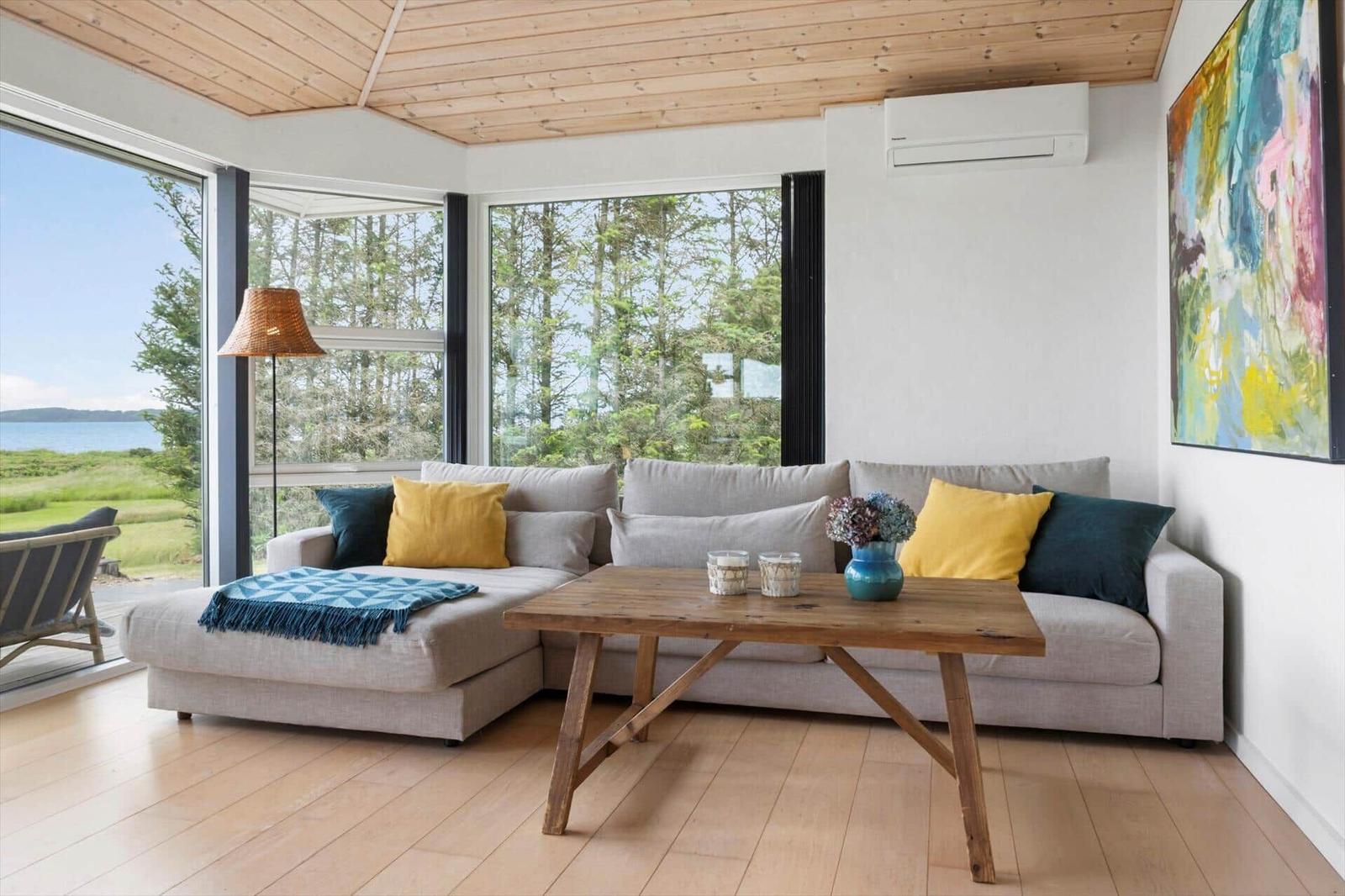 Wohnzimmer mit Sofa, Holztisch und Blick auf Wald und Wasser.