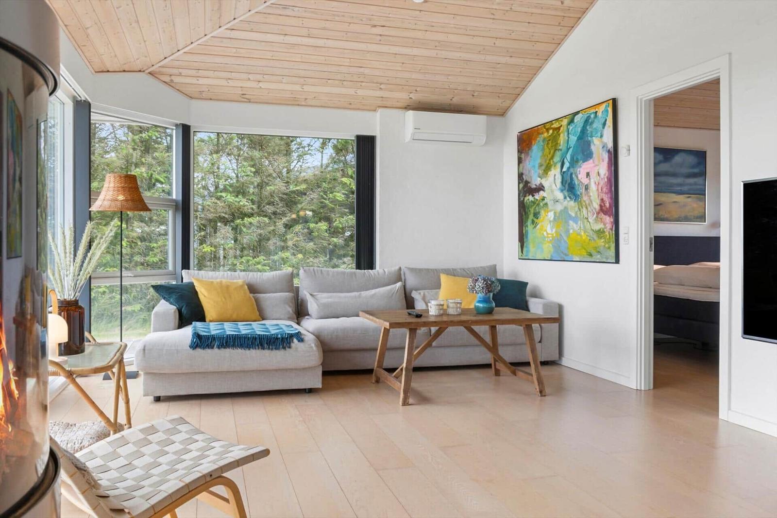 Wohnzimmer mit Sofa, Tisch und Blick auf Wald. Holzdecke und Boden. Wandbild und Kamin.