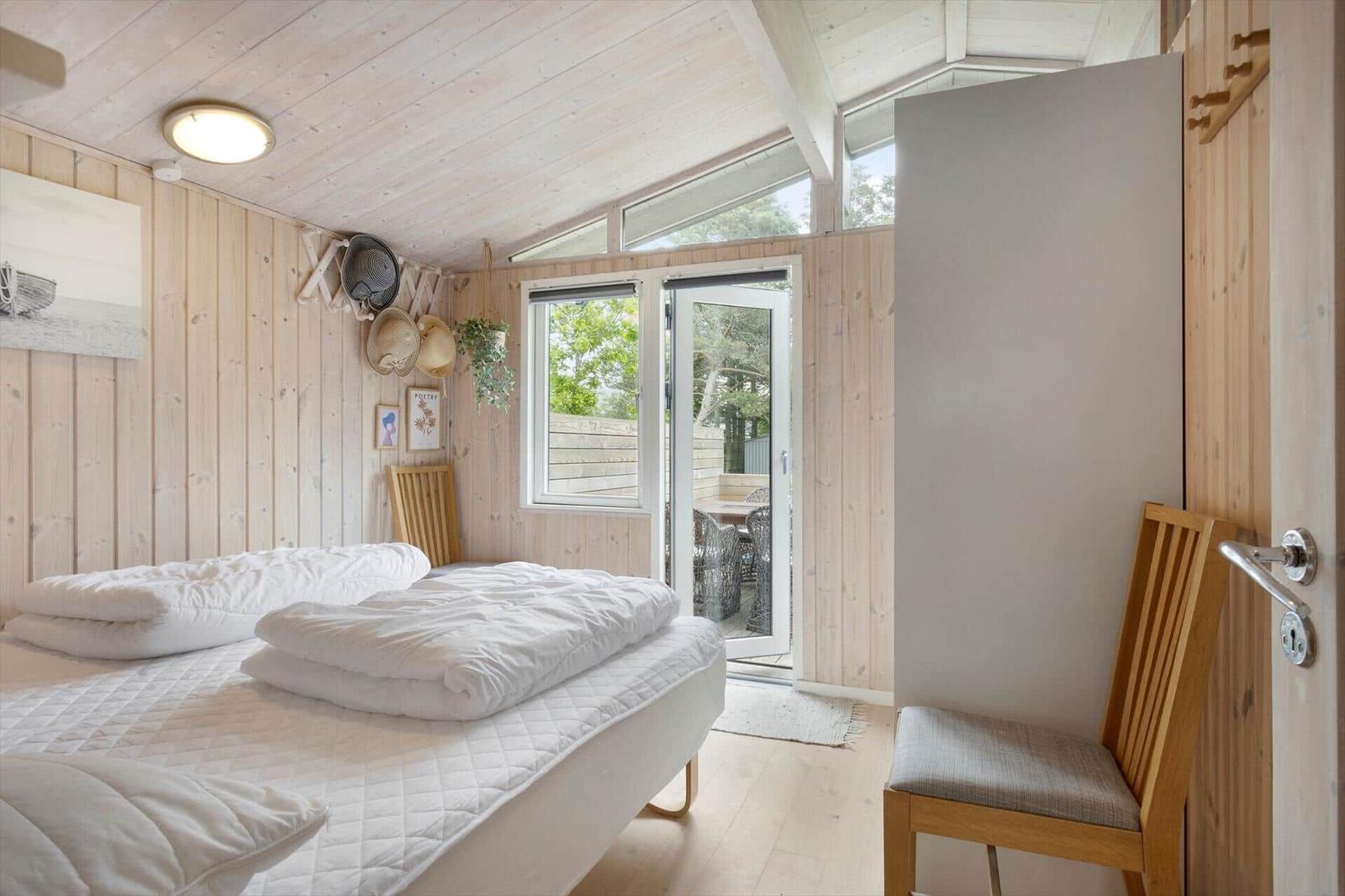 Schlafzimmer mit Bett, Holzwand und Terrassentür. Stuhl und Wanddekoration sichtbar.