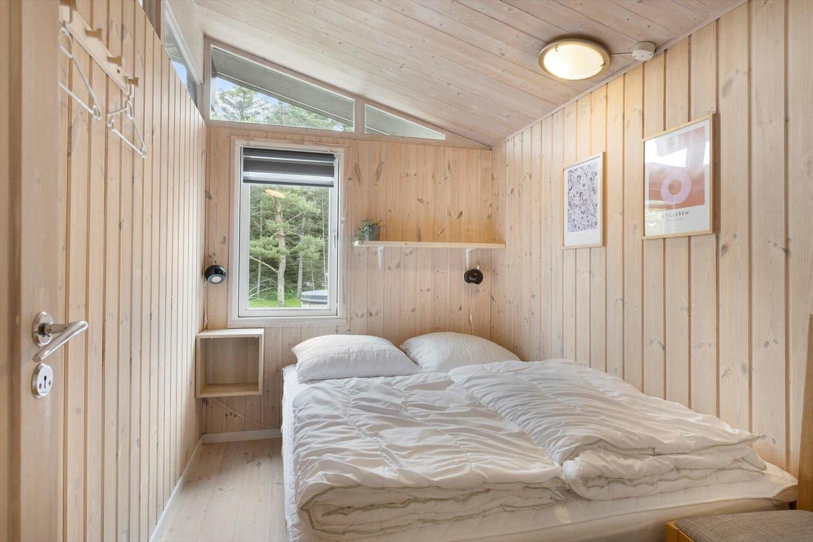Schlafzimmer mit Holzwandverkleidung, Bett und Fenster zum Blick in den Wald.