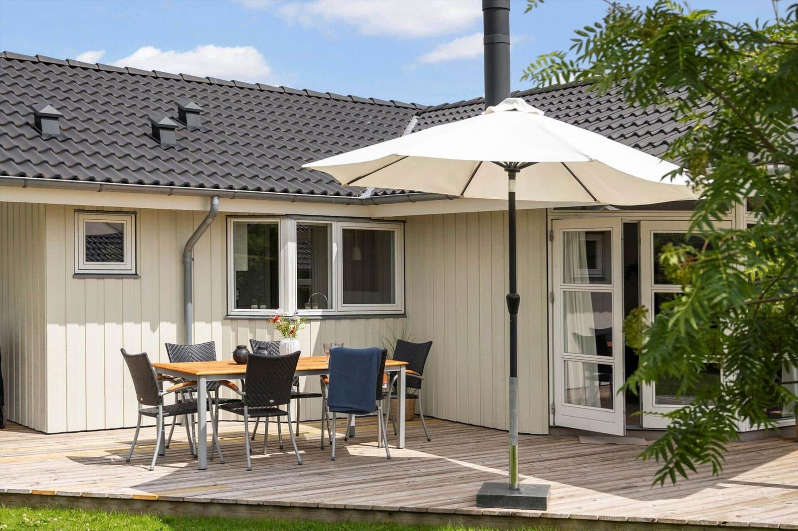 Deck with table, chairs, and umbrella in front of a house with wooden siding and roof.