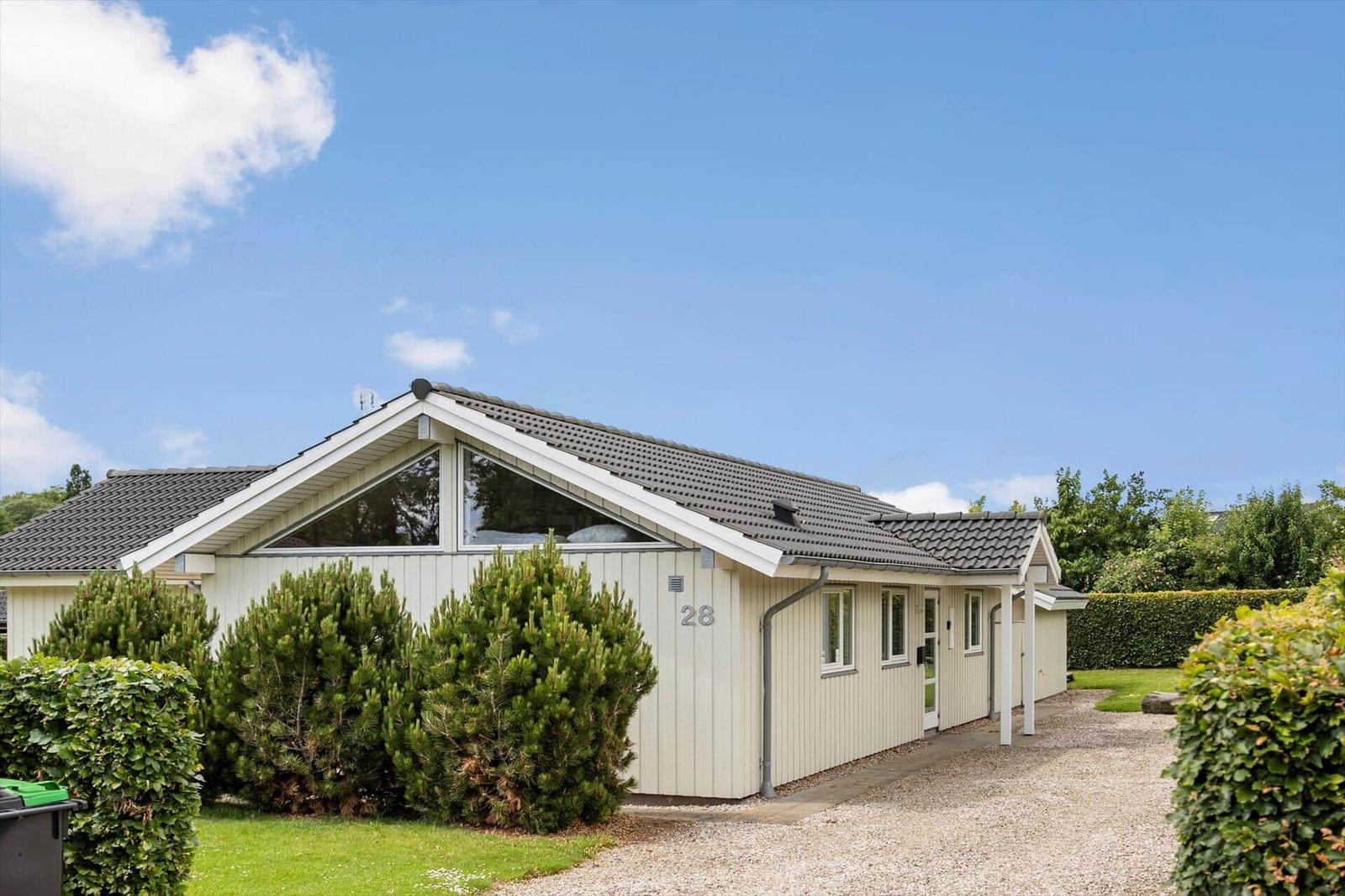A modern holiday home with light walls and gray roof. Surrounded by greenery and a gravel path.