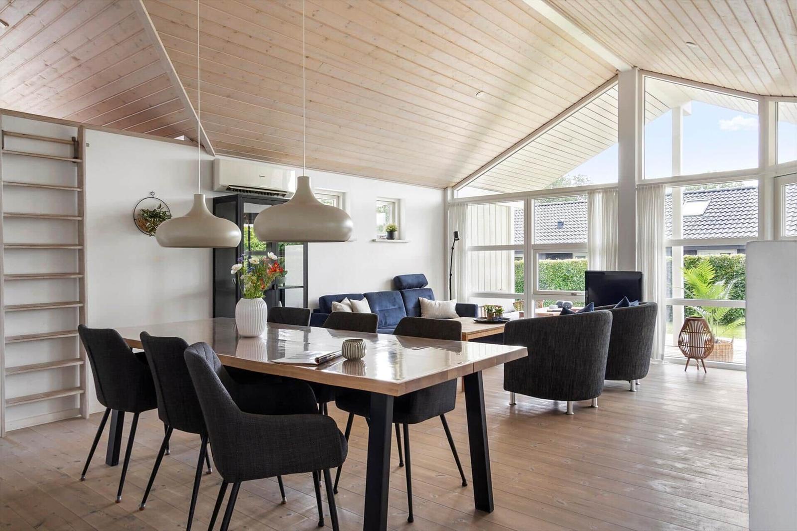Dining area with table and chairs under wooden ceiling. Living area with sofa and TV. Large window front.