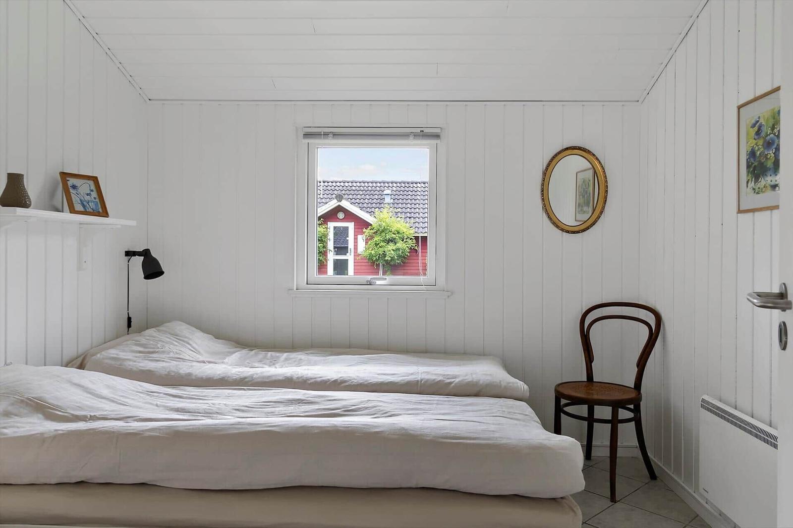 Two beds, window with view of a red house, chair and mirror on the wall.