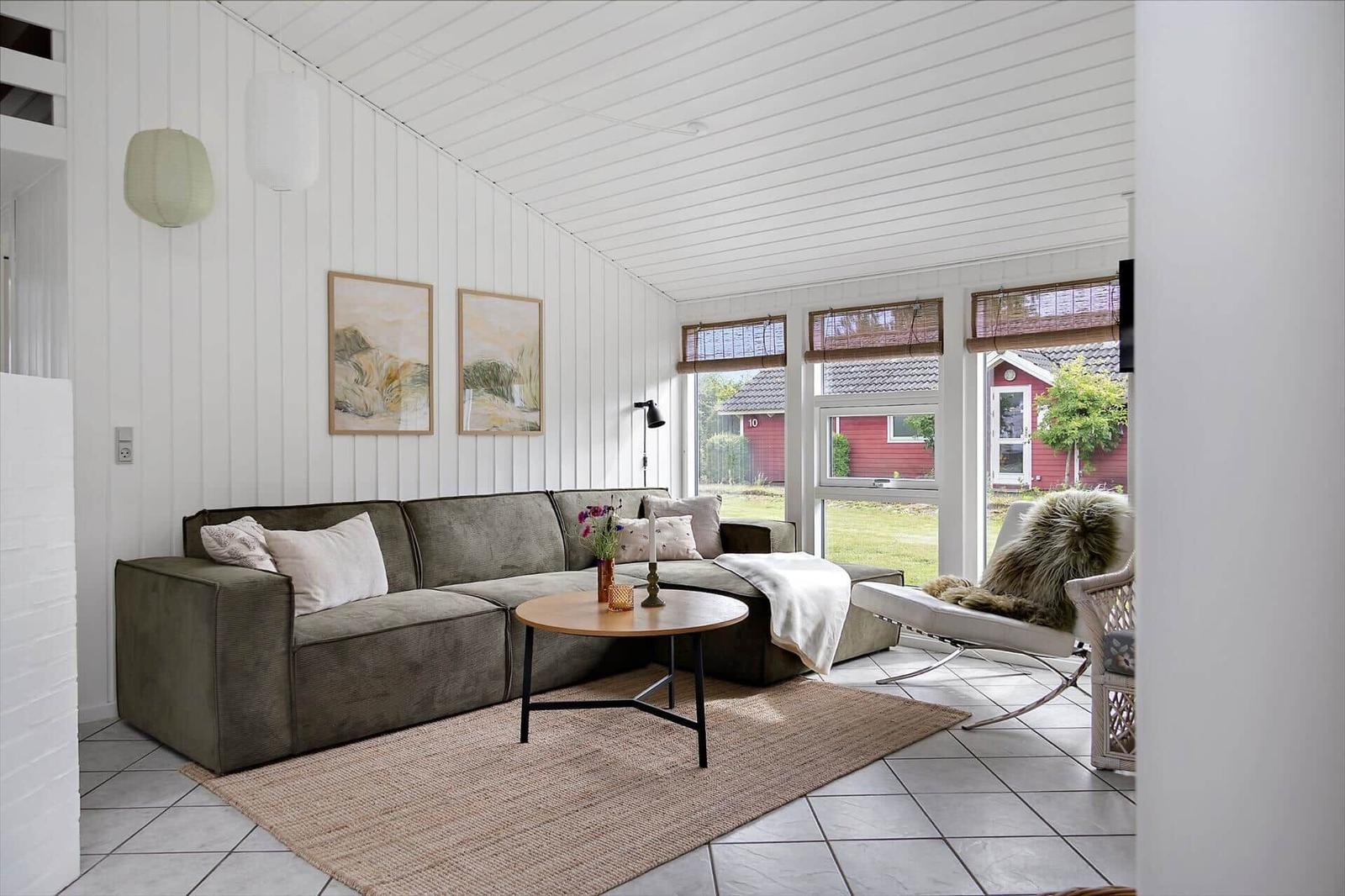 Living room with sofa, table, and chair. Windows show garden and red house.