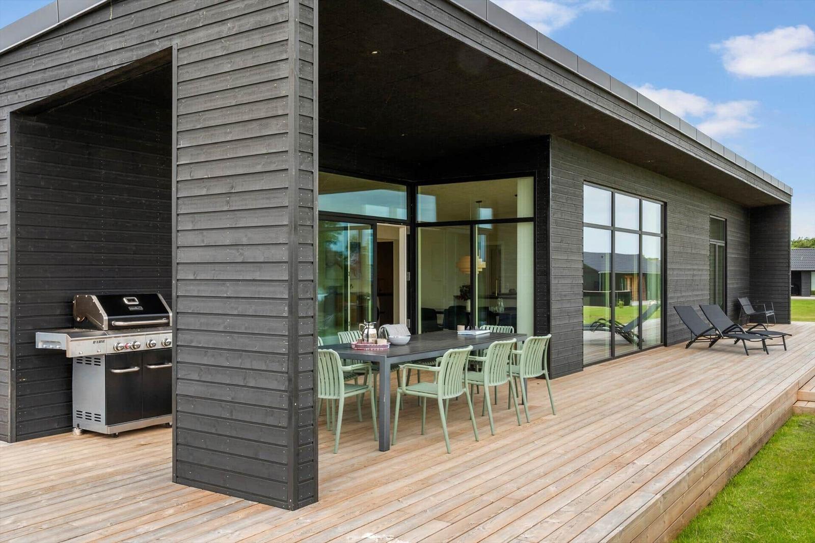 Terrasse mit Grill, Esstisch und Liegestühlen. Modernes Haus mit großem Glasbereich.
