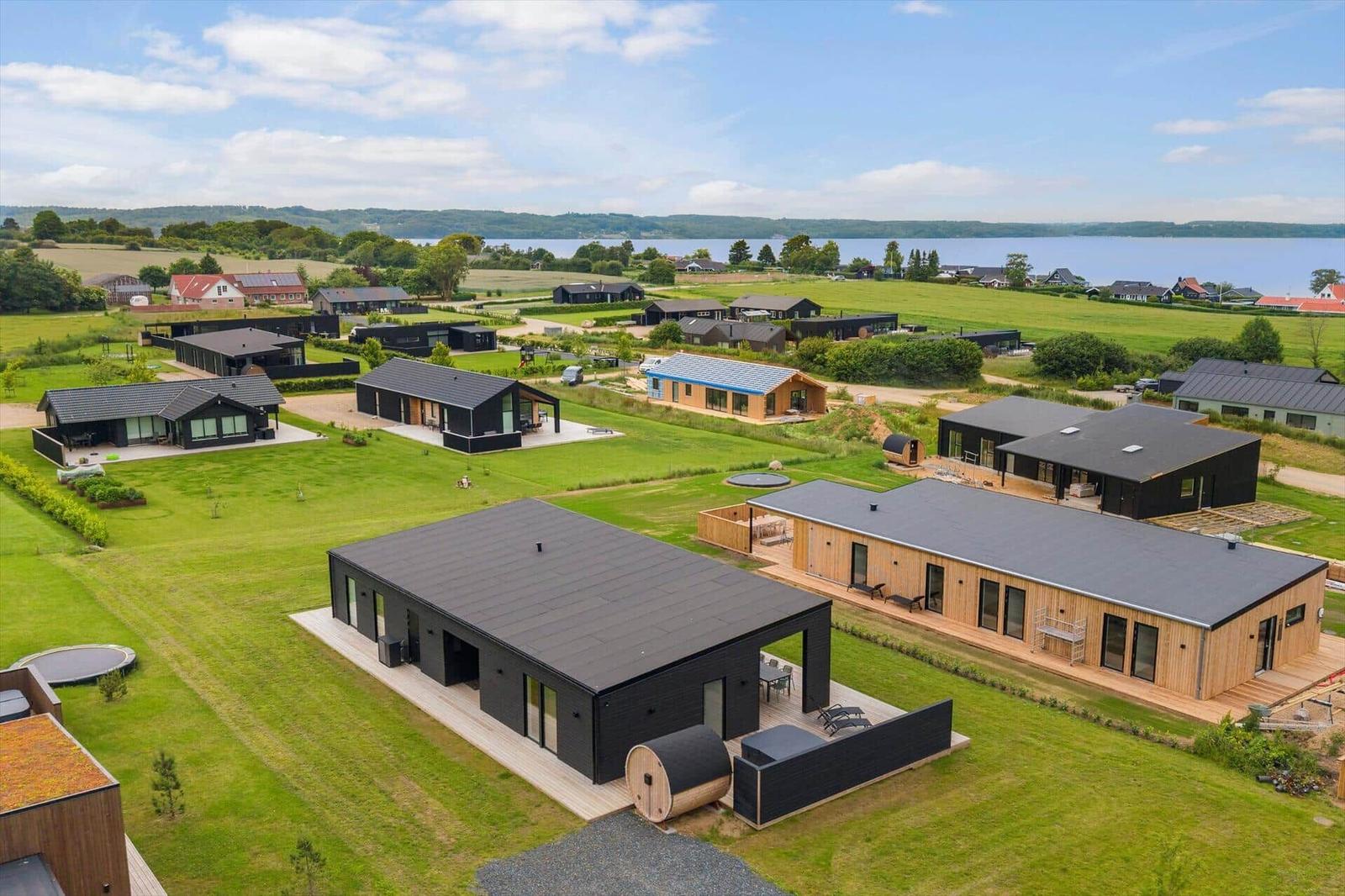 Moderne Ferienhäuser mit Holzfassade und schwarzen Dächern liegen auf grünen Wiesen am Wasser.